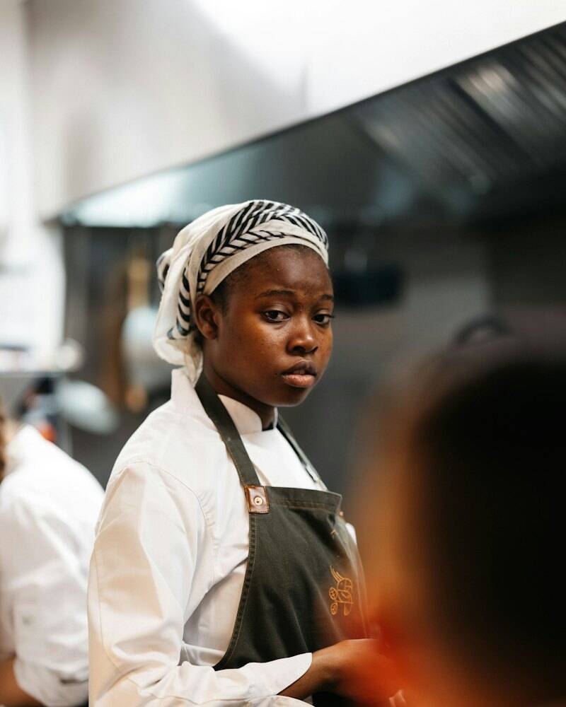 Amara Okafor in Kochjacke und Kopftuch in der Küche